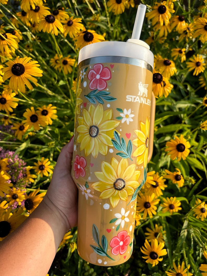 May include: A Stanley tumbler with a floral design featuring yellow sunflowers, pink and white flowers, and green leaves. The tumbler is tan with a white lid and straw. The background is a field of yellow flowers.