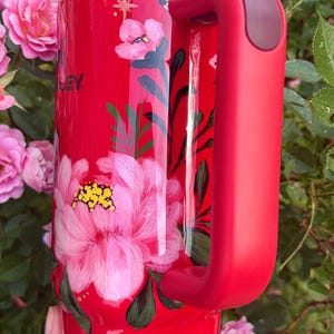 May include: A red insulated tumbler with a large handle and a clear lid featuring a white straw with red heart patterns. The exterior is decorated with hand-painted pink flowers, green leaves, and small white star accents. The word STANLEY is visible.