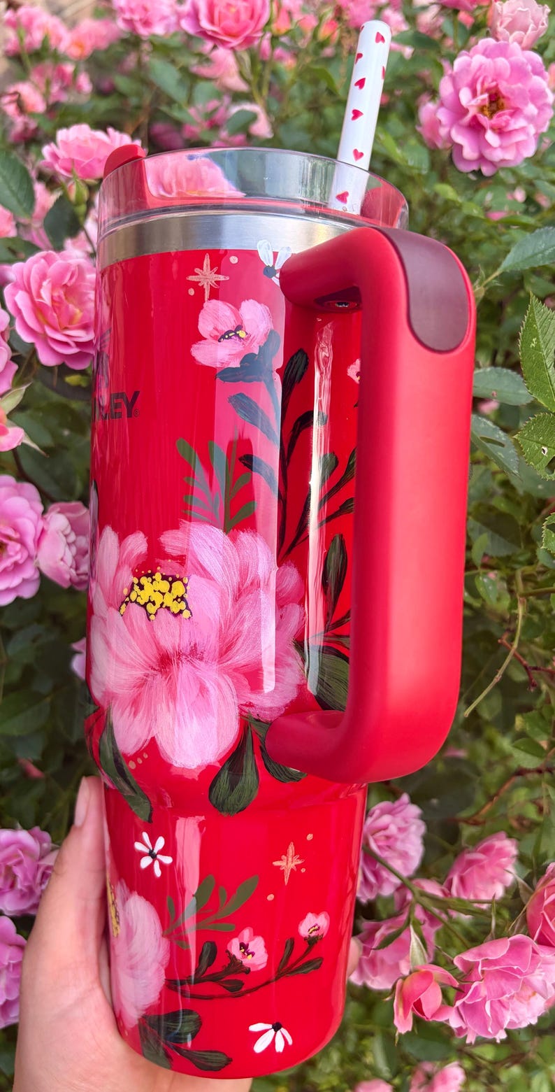 May include: A red insulated tumbler with a large handle and a clear lid featuring a white straw with red heart patterns. The exterior is decorated with hand-painted pink flowers, green leaves, and small white star accents. The word STANLEY is visible.