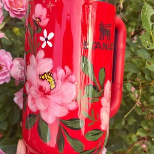 May include: A bright red Stanley tumbler featuring a hand-painted floral design with large pink peonies, green leaves, and small white star accents. The cup includes a clear lid, a red handle, and a white straw decorated with small red hearts.