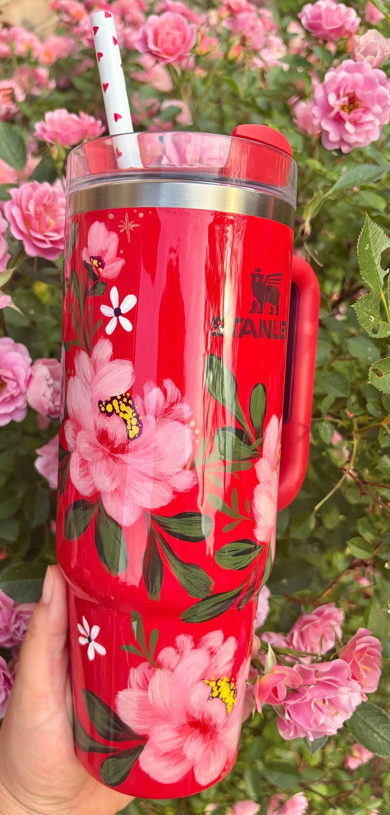 May include: A bright red Stanley tumbler featuring a hand-painted floral design with large pink peonies, green leaves, and small white star accents. The cup includes a clear lid, a red handle, and a white straw decorated with small red hearts.