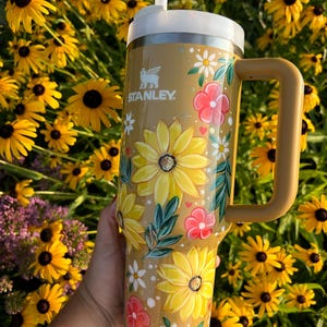 May include: A tan Stanley tumbler with a white lid and handle, adorned with hand-painted yellow sunflowers, pink flowers, and white daisies. The tumbler has the Stanley logo and a white straw.