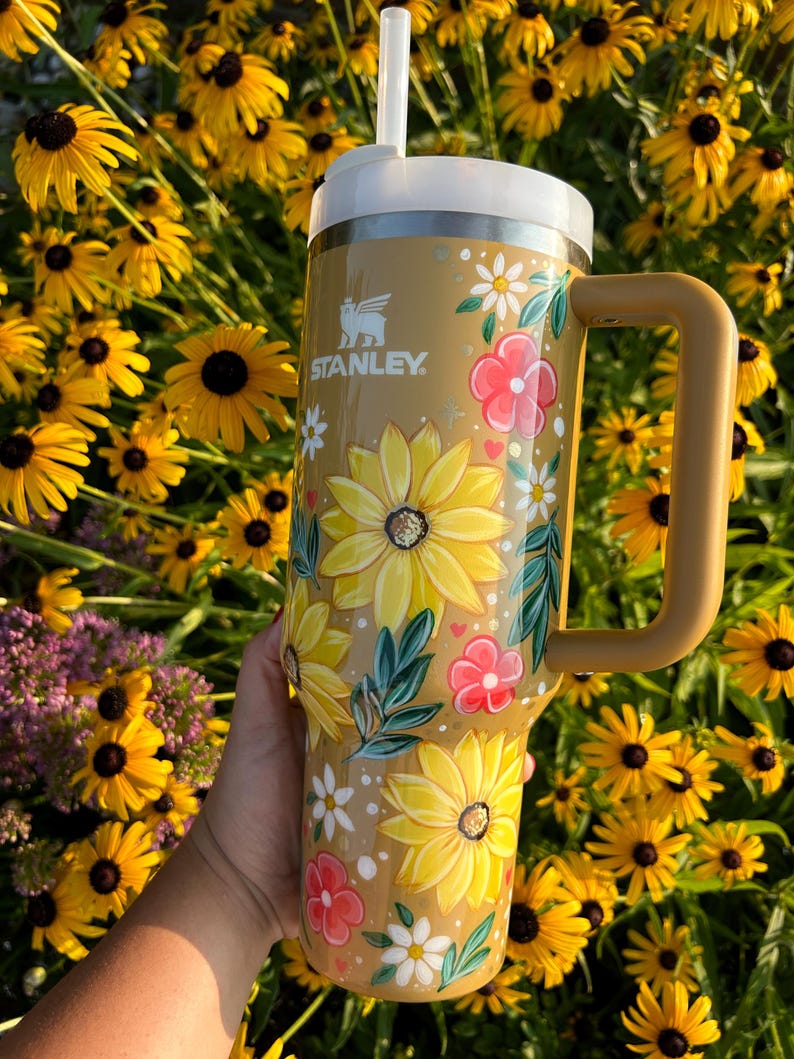 May include: A tan Stanley tumbler with a white lid and handle, adorned with hand-painted yellow sunflowers, pink flowers, and white daisies. The tumbler has the Stanley logo and a white straw.