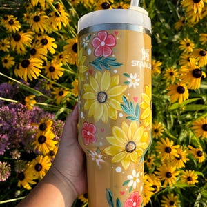 May include: A tan Stanley tumbler with a white lid and straw, adorned with hand-painted yellow sunflowers, red and white flowers, and green leaves. The tumbler is held in front of a backdrop of yellow flowers.