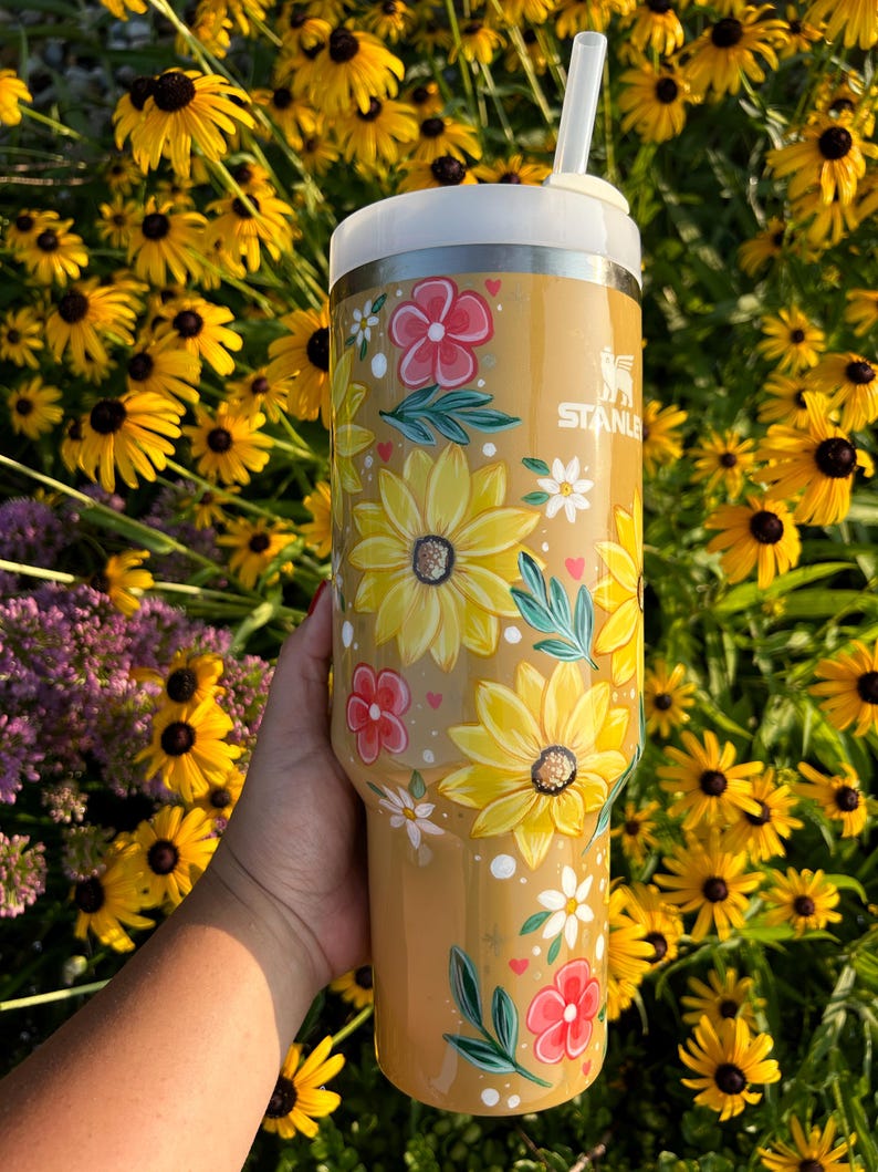 May include: A tan Stanley tumbler with a white lid and straw, adorned with hand-painted yellow sunflowers, red and white flowers, and green leaves. The tumbler is held in front of a backdrop of yellow flowers.