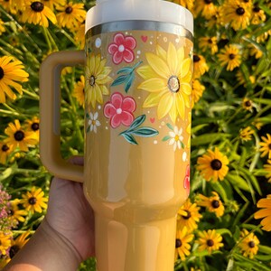 May include: A yellow insulated tumbler with a white lid and handle. The tumbler is decorated with hand-painted yellow sunflowers, red flowers, and green leaves. The tumbler has a straw.