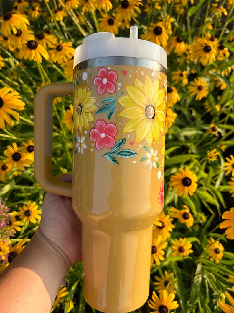 May include: A yellow insulated tumbler with a white lid and handle. The tumbler is decorated with hand-painted yellow sunflowers, red flowers, and green leaves. The tumbler has a straw.