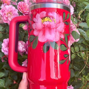 May include: A bright red insulated tumbler with a handle, featuring a hand-painted pink peony design with green leaves. The tumbler includes a clear lid and a white straw decorated with small red hearts, set against a vibrant floral background.