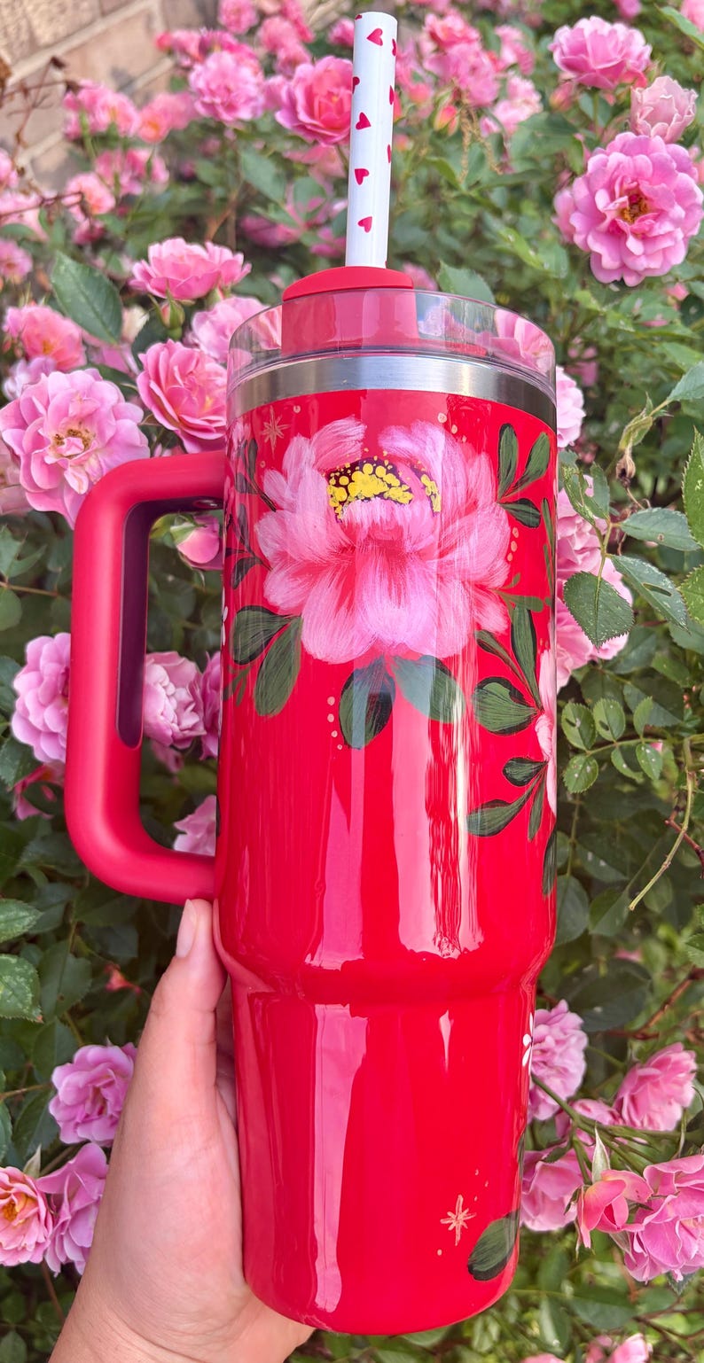 May include: A bright red insulated tumbler with a handle, featuring a hand-painted pink peony design with green leaves. The tumbler includes a clear lid and a white straw decorated with small red hearts, set against a vibrant floral background.