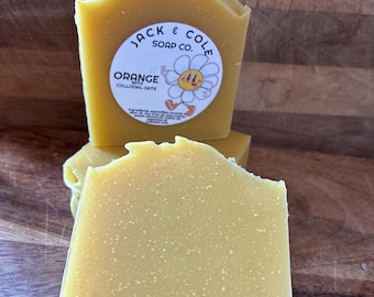 May include: Close-up of several bars of orange-colored soap. The soap has a rough, textured surface and a label that reads "Jack & Cole Soap Co." with the words "Orange with Colloidal Oats" and a daisy graphic.