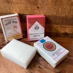 May include: Three bars of soap packaged in vintage-style boxes. The boxes are red, white, and green with gold lettering. The soap bars are white and are labeled "Candy Cigarette Scented Soap Bar" and "Lucky Soap".