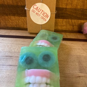 May include: Green soap bars with a face design featuring large blue eyes and a set of pink-gums teeth. A small brown box with a "CAUTION DO NOT OPEN" sticker is tied with twine.