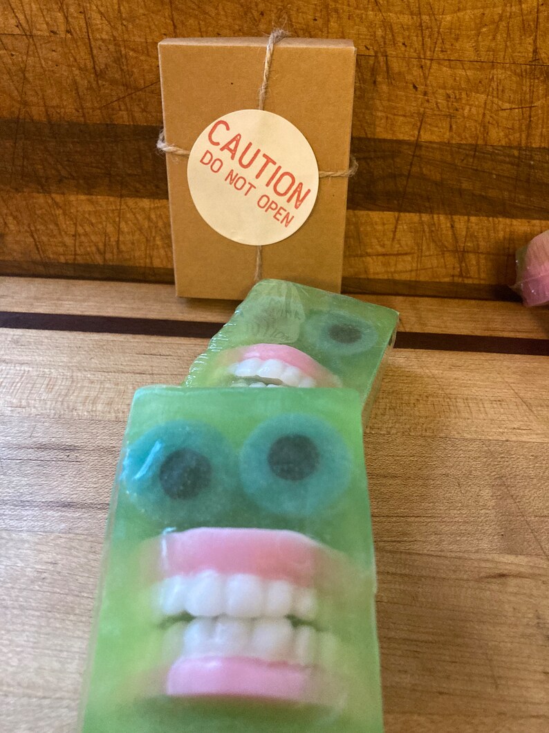 May include: Green soap bars with a face design featuring large blue eyes and a set of pink-gums teeth. A small brown box with a "CAUTION DO NOT OPEN" sticker is tied with twine.