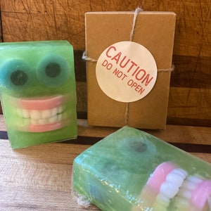 May include: Green soap bars with a cartoon face design, featuring large eyes and a smiling mouth with white teeth and pink gums. A brown box with a "CAUTION DO NOT OPEN" sticker is also present.