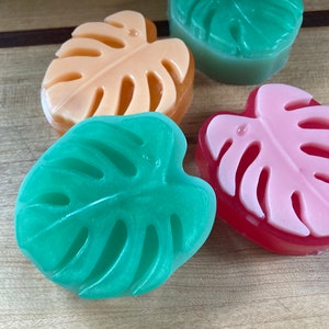 May include: Four handmade soap bars shaped like monstera leaves. The soaps are in shades of green, pink, and orange.