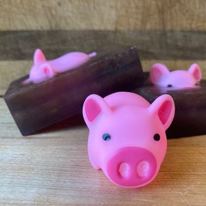 May include: Three pink pig-shaped soaps on brown soap bars. The pig soaps have black eyes and pink snouts.