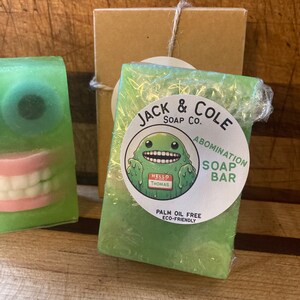 May include: Green soap bars with monster-themed designs. One bar features a large eye, the other a set of teeth. A wrapped bar has a label that reads "Jack & Cole Soap Co. ABOMINATION SOAP BAR" with a cartoon monster and "HELLO THOMAS".