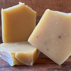 May include: Three bars of handmade soap in shades of yellow and tan are arranged on a wooden surface. The soap has a rustic, textured appearance with visible imperfections. The bars are cut into irregular shapes, suggesting a natural, artisanal product.