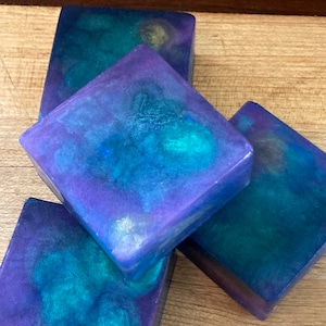 May include: Four square bars of handmade soap with a galaxy-inspired design. The soap is a mix of purple, blue, and green colors with swirls and flecks of gold.