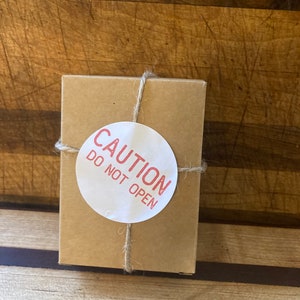 May include: A small, square, brown cardboard box tied with twine. A white circular sticker with the red text "CAUTION DO NOT OPEN" is affixed to the box. The box is on a wooden surface.