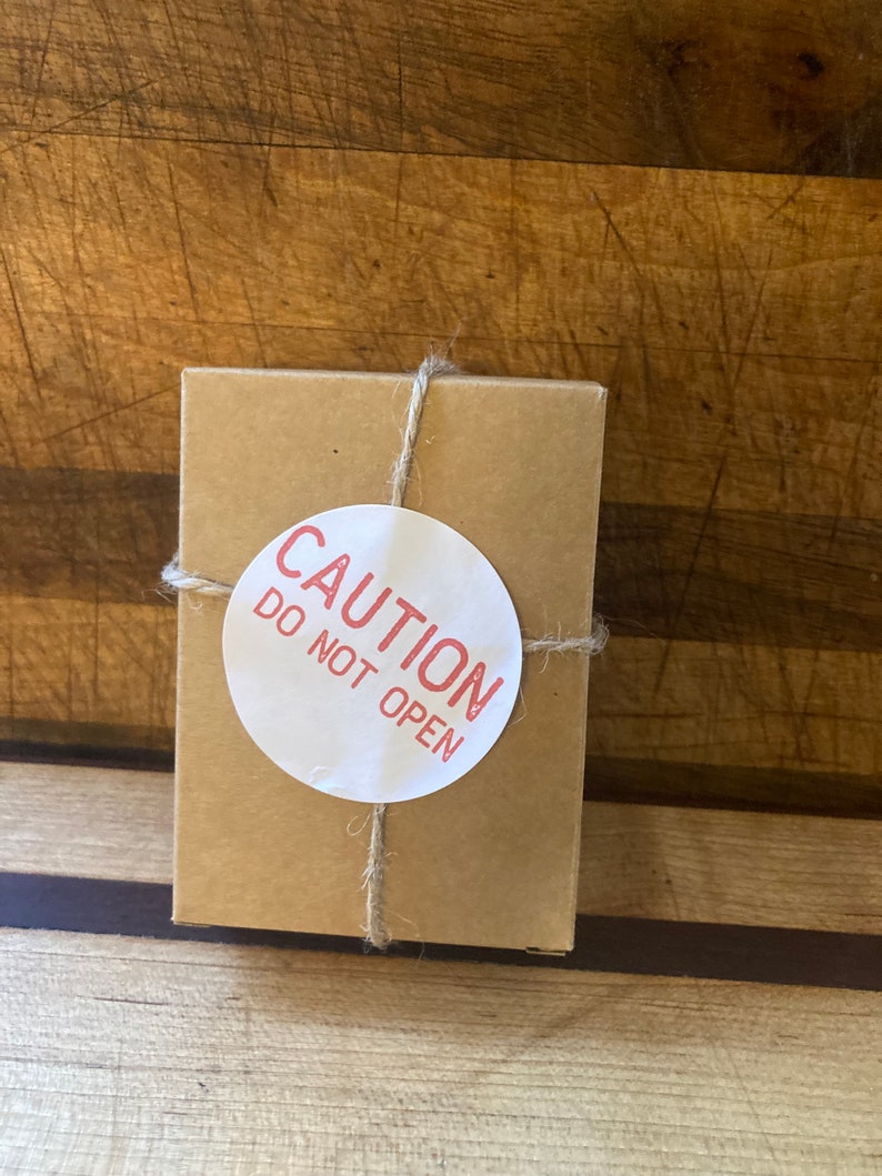 May include: A small, square, brown cardboard box tied with twine. A white circular sticker with the red text "CAUTION DO NOT OPEN" is affixed to the box. The box is on a wooden surface.