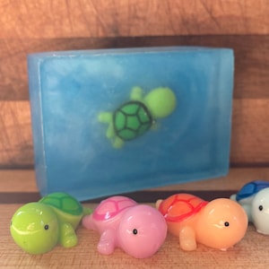 May include: Five small turtle figurines, each a different color: green, pink, orange, blue, and light blue. The turtles are arranged in a row on a wooden surface. A blue soap bar with a green turtle design is in the background.