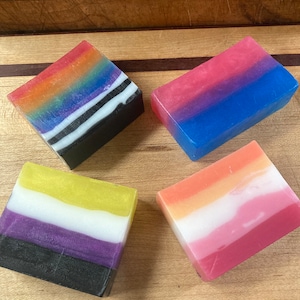 May include: Four bars of soap with different color combinations, each representing a different pride flag. The flags are rainbow, bisexual, non-binary, and transgender.