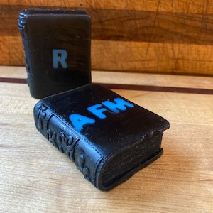 May include: Two small black books with decorative covers. The book on the left has the letter "R" on the front. The book on the right has the letters "AFM" on the front.