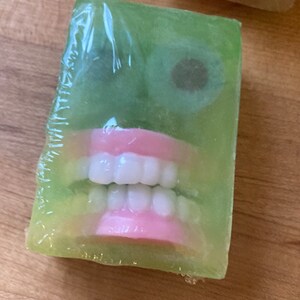 May include: A green bar of soap with a face design. The soap features white teeth, pink gums, and two dark brown eye shapes. The soap is wrapped in clear plastic.