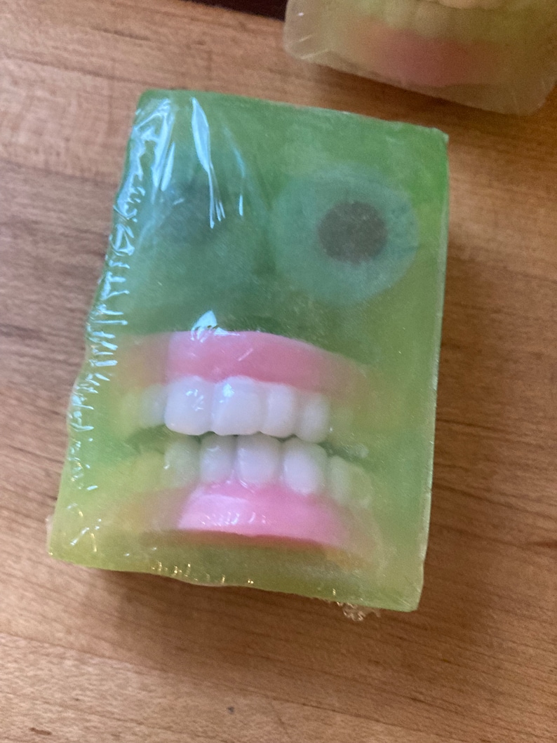May include: A green bar of soap with a face design. The soap features white teeth, pink gums, and two dark brown eye shapes. The soap is wrapped in clear plastic.