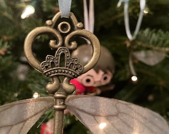 May include: A gold-colored key with a crown design and intricate details, adorned with delicate white wings. The key is hanging from a blue ribbon on a Christmas tree.