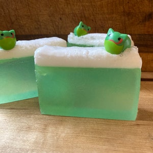 May include: Three bars of green and white soap with a frog on top of each bar. The soap is on a wooden surface.