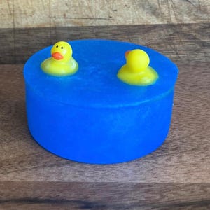 May include: A blue soap bar with two yellow rubber duckies on top. The duckies are floating in the soap, which is shaped like a pool.