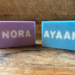 May include: Two rectangular bars of soap. One is lavender with the name "NORA" in white letters. The other is light blue with the name "AYAAN" in white letters. The soaps are displayed on a wooden surface.