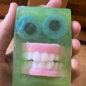 May include: A novelty soap bar with a green translucent body. The soap features two large, round, teal-colored eyes with black pupils and a set of pink-gums and white teeth. The soap is held in a hand.