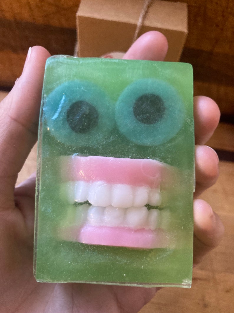 May include: A novelty soap bar with a green translucent body. The soap features two large, round, teal-colored eyes with black pupils and a set of pink-gums and white teeth. The soap is held in a hand.