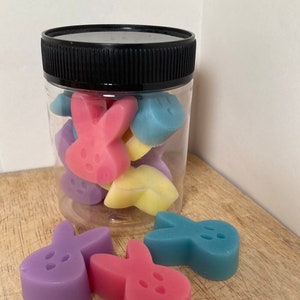 May include: A clear glass jar with a black lid filled with pastel colored bunny shaped wax melts.  The jar is sitting on a wooden surface.  There are three additional bunny shaped wax melts in front of the jar.