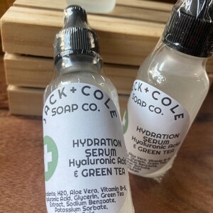 May include: Two clear glass bottles with black caps and white labels. The labels read "CK + COLE SOAP CO." and "HYDRATION SERUM Hyaluronic Acid & GREEN TEA". The bottles are sitting on a wooden surface.