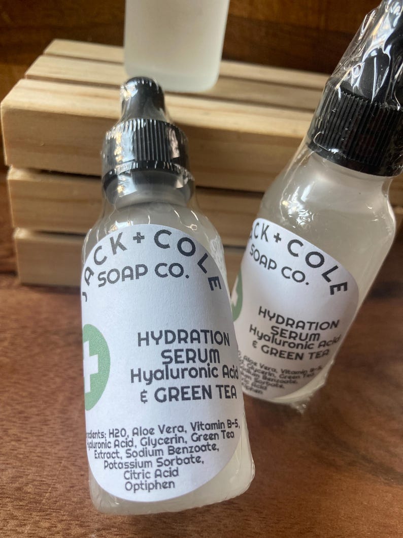 May include: Two clear glass bottles with black caps and white labels. The labels read "CK + COLE SOAP CO." and "HYDRATION SERUM Hyaluronic Acid & GREEN TEA". The bottles are sitting on a wooden surface.