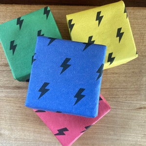 May include: Four small gift boxes stacked on top of each other. The boxes are wrapped in different colors: green, yellow, blue, and pink. Each box has a black lightning bolt pattern.