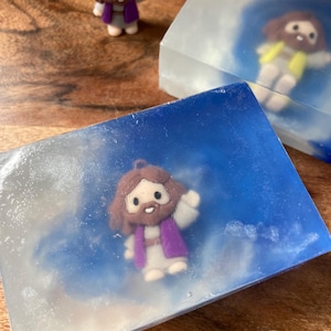 May include: Handmade soap bars with a religious theme. Each bar features a small figurine of Jesus Christ, set against a blue and white cloudy background. The soap bars are rectangular and have a clear, translucent appearance.