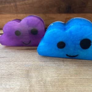 May include: Two cloud-shaped decorative items. One is purple, the other is blue. Both have black eyes and smiling mouths. They are placed on a wooden surface. The items appear to be made of a soft material.
