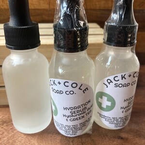 May include: Three clear glass bottles with black dropper tops and white labels. The labels have green accents and text that reads "Jack + Cole Soap Co." and "Hydration Serum Hyaluronic Acid & Green Tea".