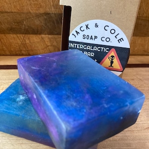 May include: Two bars of soap with a galaxy-themed design. The soap is packaged in a cardboard box with the text "Jack & Cole Soap Co." and "Intergalactic Soap Bar" printed on it. The box also features a warning sign with an alien inside.