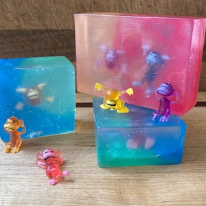 May include: Three clear soap bars with different colored layers. Each bar has a small plastic monkey toy inside. The monkeys are yellow, purple, and pink.