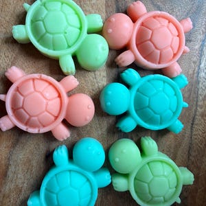 May include: Six colorful turtle-shaped soaps in shades of green, pink, and blue. The soaps have a detailed shell design and are arranged on a wooden surface. The turtles are a fun and decorative item.