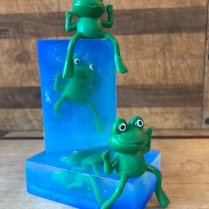 May include: Two green frog figurines sitting on top of blue soap bars. The frogs have large eyes and are posed with their arms raised. The soap bars are translucent and have a light blue color.