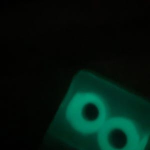 May include: A glowing green, rectangular object with two large, circular eye shapes. The eyes have black pupils and a bright green outline, creating a spooky effect against the dark background. The object appears to be made of a translucent material.
