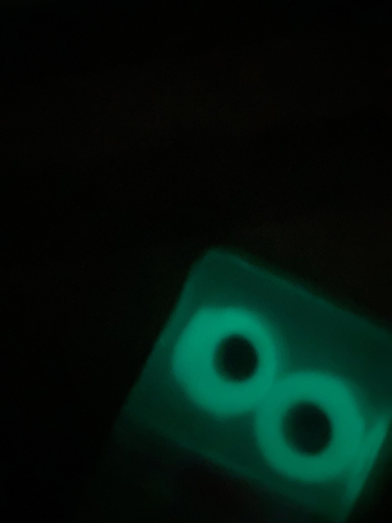 May include: A glowing green, rectangular object with two large, circular eye shapes. The eyes have black pupils and a bright green outline, creating a spooky effect against the dark background. The object appears to be made of a translucent material.
