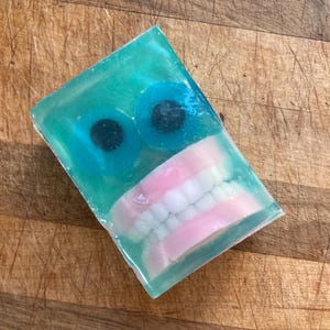 May include: A rectangular bar of soap with a teal translucent base. Two blue, round candies with black centers are set near the top. A set of pink-based dentures is embedded in the soap.